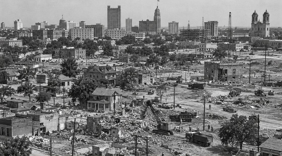 Illustrated reconstruction: Black and white view of the clearance: rubble, dump trucks and a crane among the few houses still standing, downtown behind.